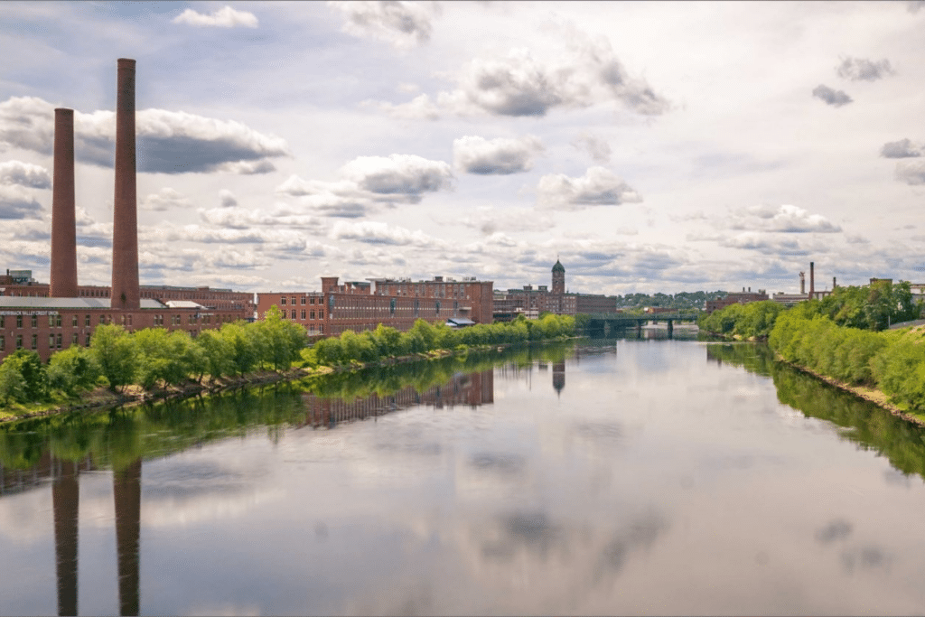 A serene Merrimack River scene with reflections of clouds and industrial buildings. Two tall chimneys rise on the left, while lush green trees line both sides of the river. A distant bridge is visible against the sky filled with fluffy clouds.