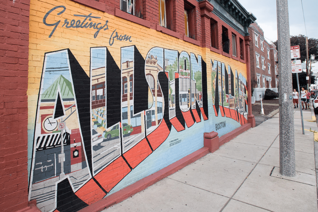 Allston MA | Allston Village mural in Boston – "Vibrant 'Greetings from Allston Village' mural on a brick wall in Boston, showcasing the neighborhood's eclectic energy, street art culture, and urban charm. | The Charley Apartments Boston MA